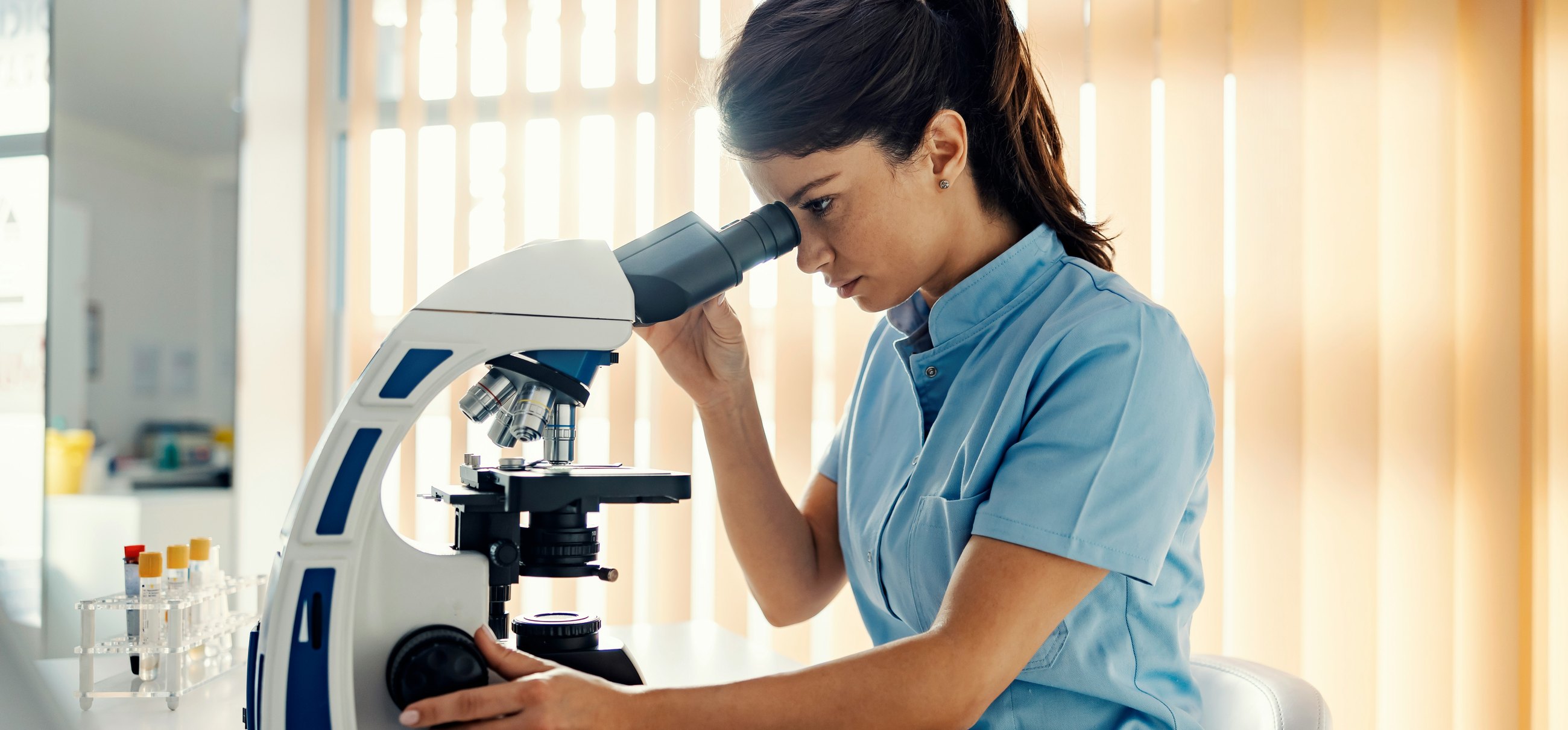 A laboratory scientist at a microscope beside a rack of sample tubes