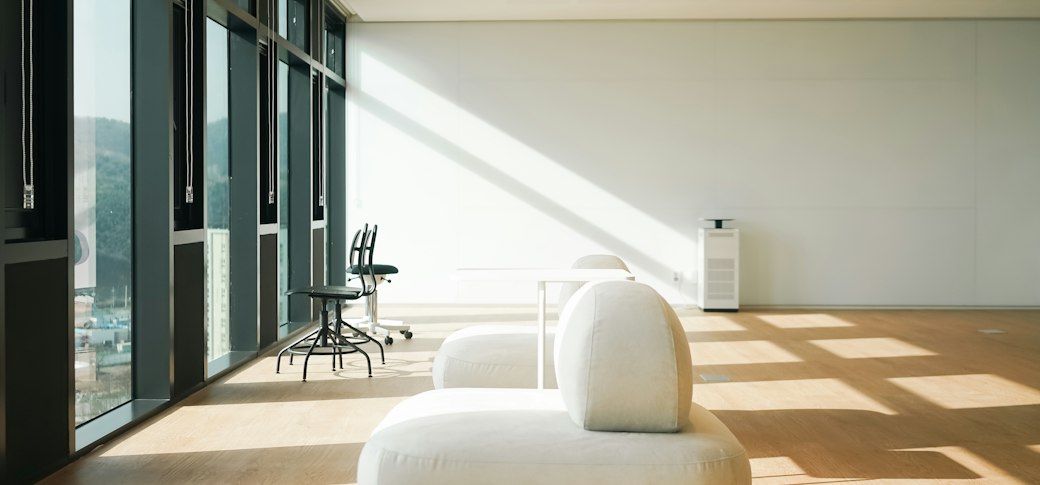 A near-empty white room with a cream curved sofa, pale oak floor and daylight raking diagonally across a blank wall.