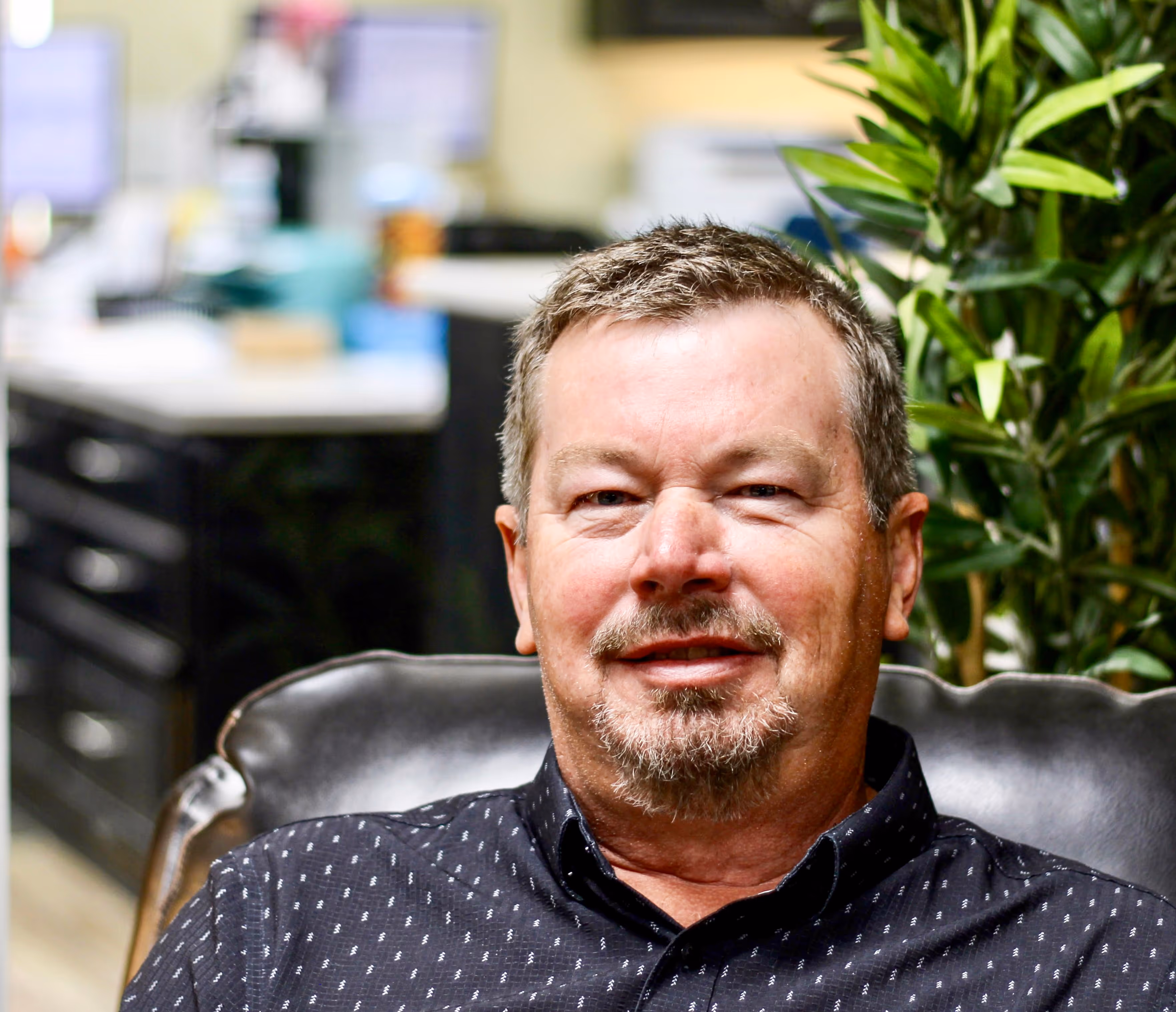 a pleasant-looking man with short hair and a salt-and-pepper beard, wearing a dark grey shirt, relaxes in a leather chair inside a professional space