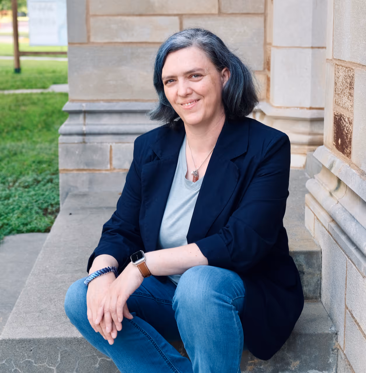a woman with dark hair in a dark jacket and jeans sits on a concrete step, smiling at the camera