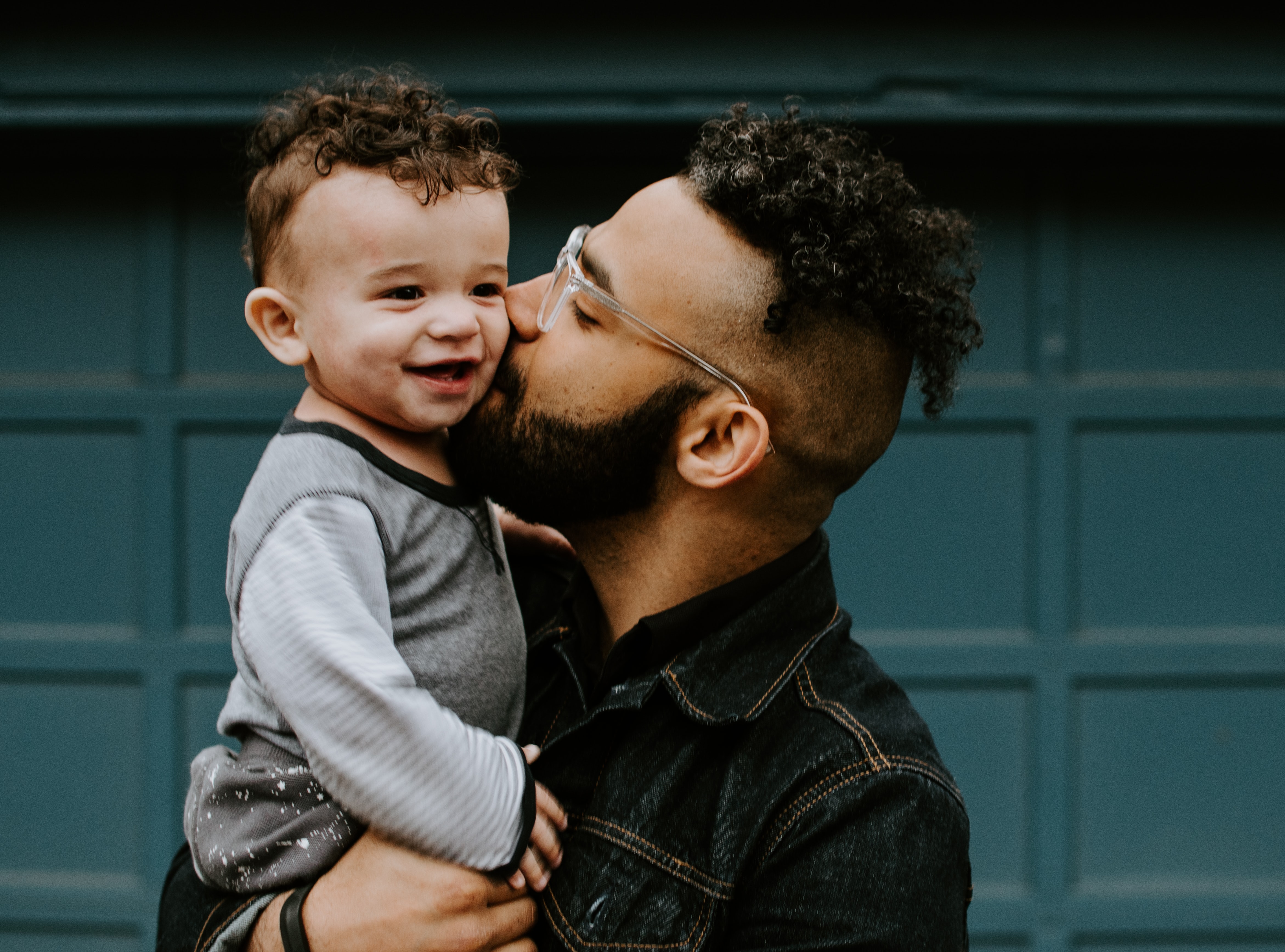 Smiling baby being kissed by father in denim jacket against teal background