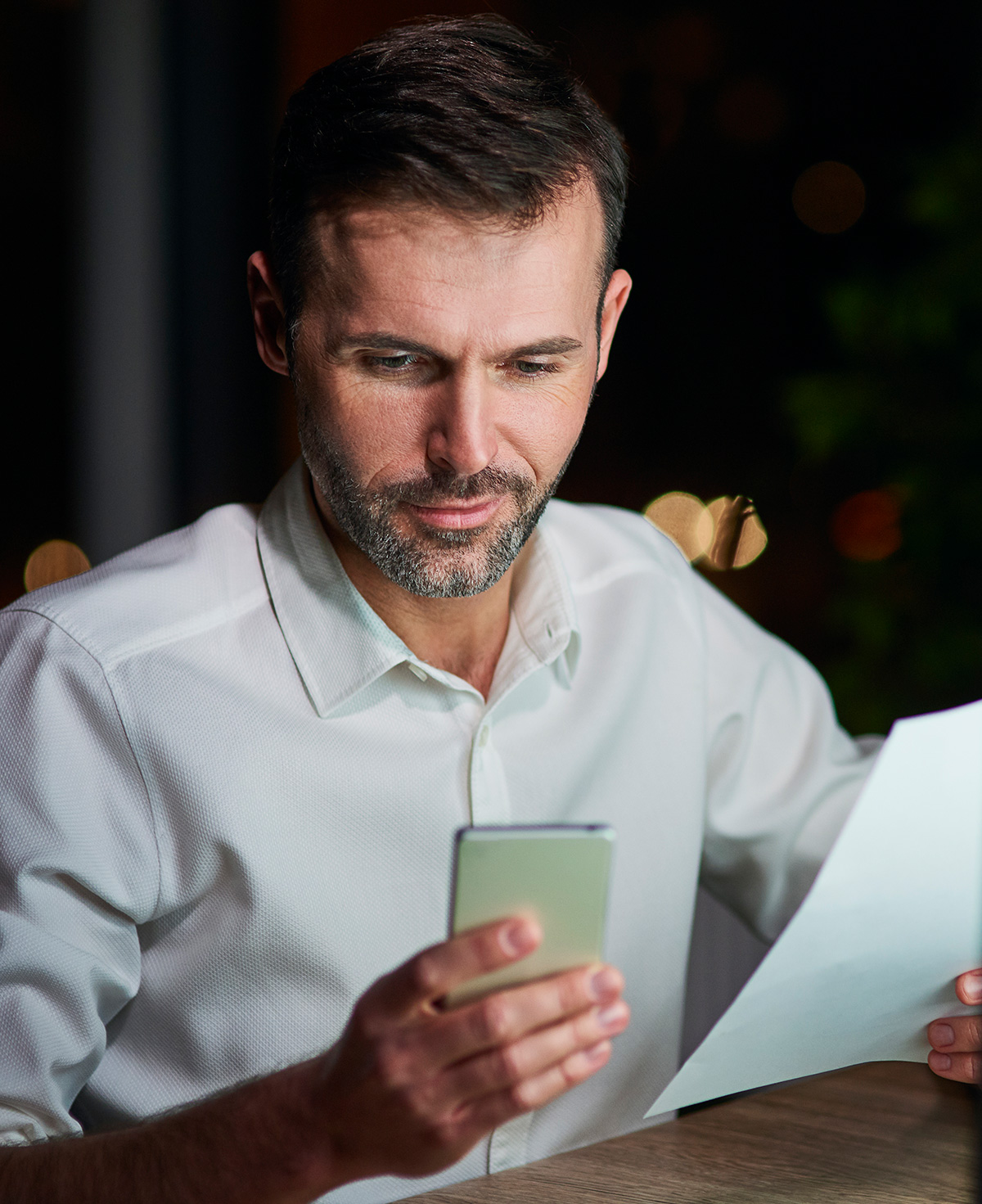 Professional man checking phone and documents at desk in low light