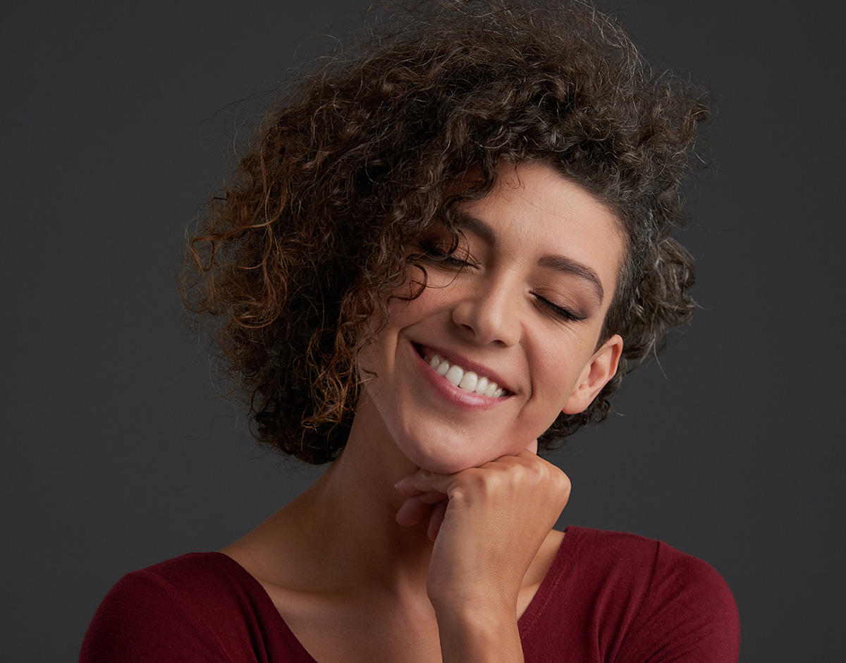 Woman with curly hair smiling with eyes closed against dark background