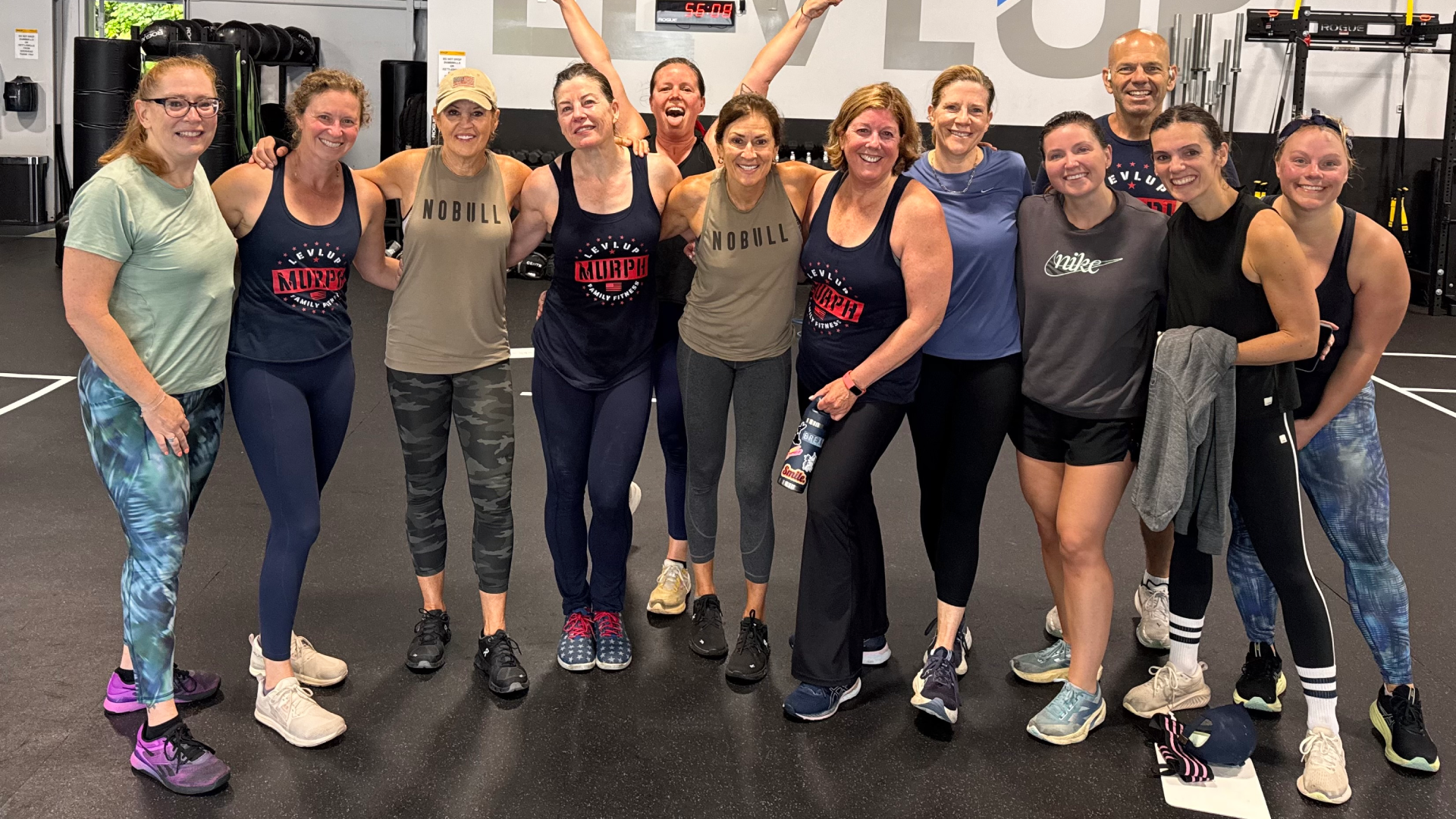 Group workout photo of smiling people in athletic gear at a gym, with "Level Up" logo on the wall. The mood is energetic and cheerful.