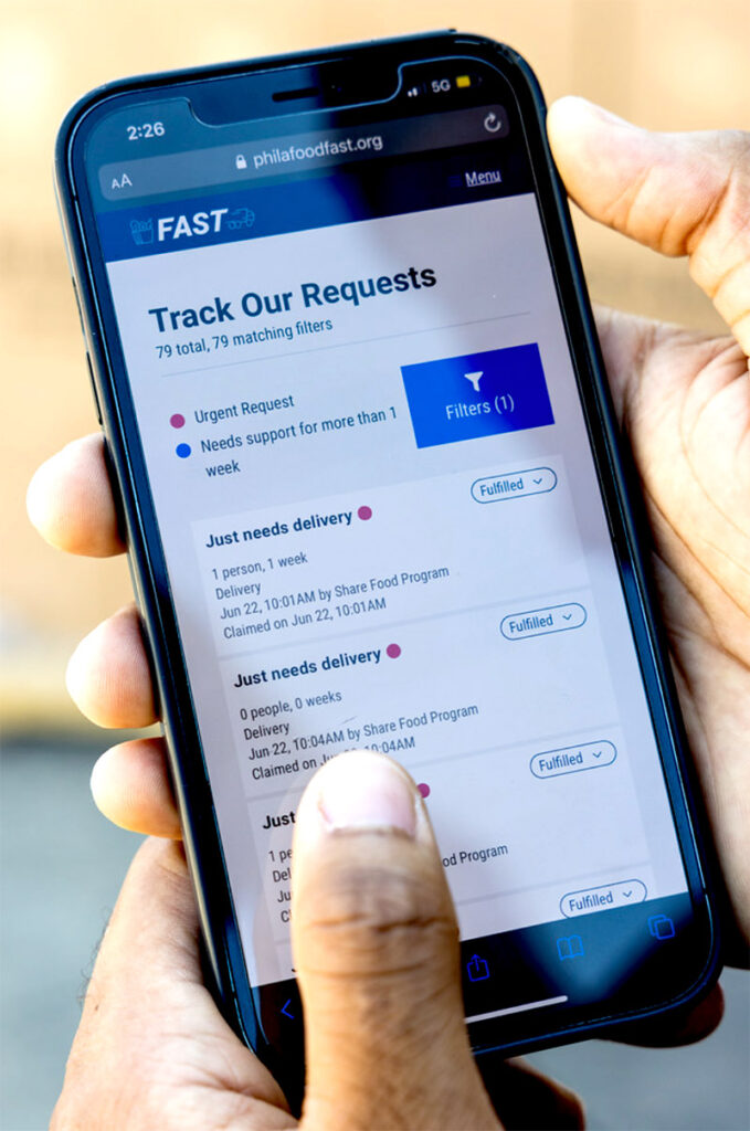 A person holds a phone in their hand. On the screen you can see a mobile app for fulfilling requests for food from food-insecure patients.