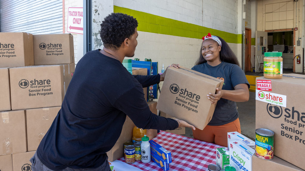 A delivery driver takes a box labeled "Share" from a woman. There are more boxes piled around them.