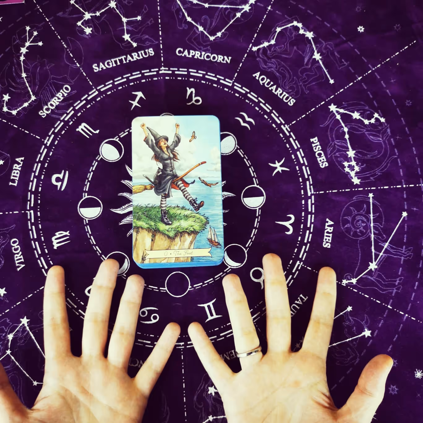 The Fool card from the Everyday Witch Tarot laid on an astrology cloth, with open hands channeling its healing energy.