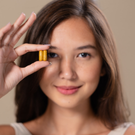 A woman holds a yellow capsule pill up to her right eye, smiling softly at the camera against a neutral background.