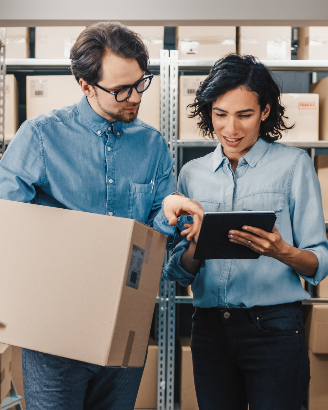 Two people stand in a warehouse surrounded by boxes, one holding a tablet while discussing logistics.