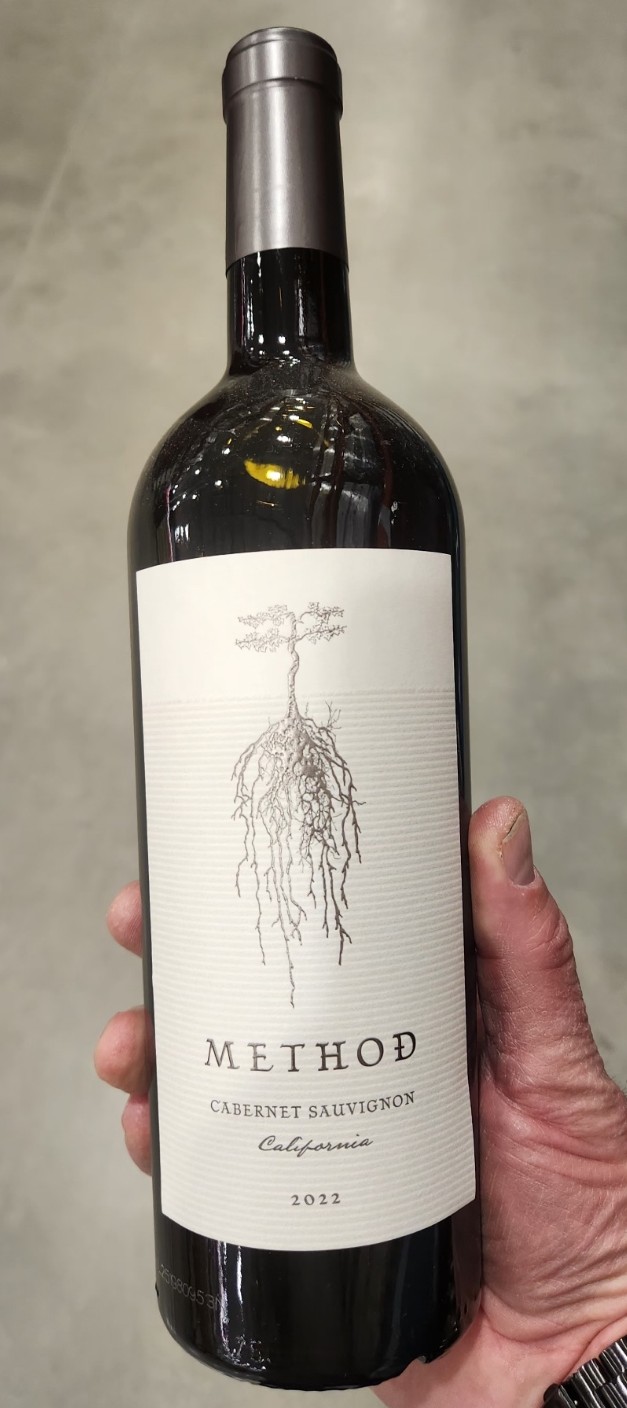 Jeff Kretschmar holding a bottle of 2022 Method Cabernet Sauvignon California in a Costco-style warehouse aisle, showcasing a great value Cabernet find.