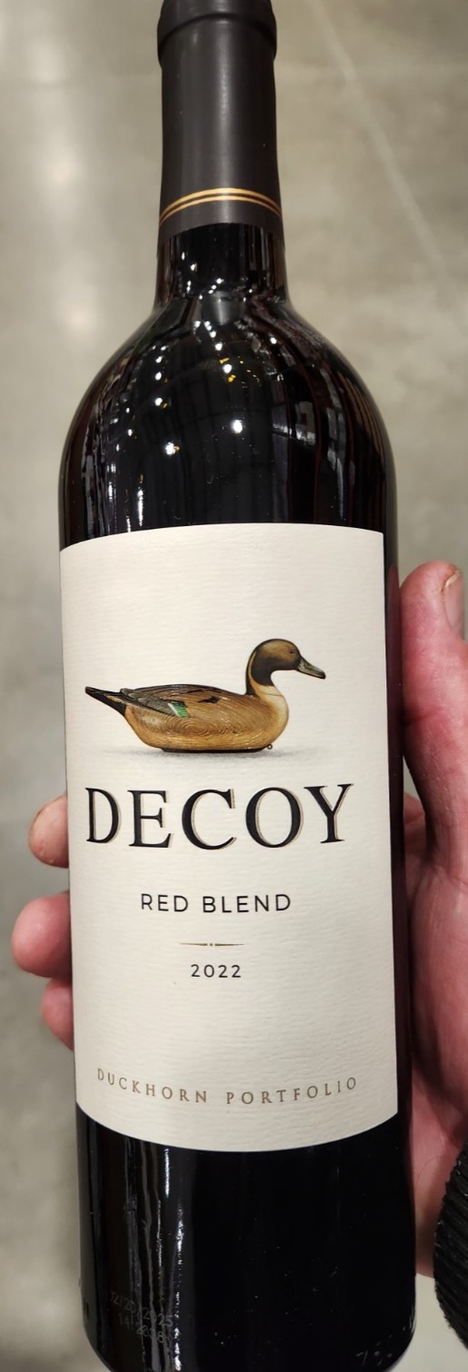 Two bottles of Decoy Red Wine on a grocery shelf above a price tag showing $29.99 regular, $20.49 sale, and $18.44 with 6+ bottles; Costco sign shows $15.89 for Decoy Red Wine.