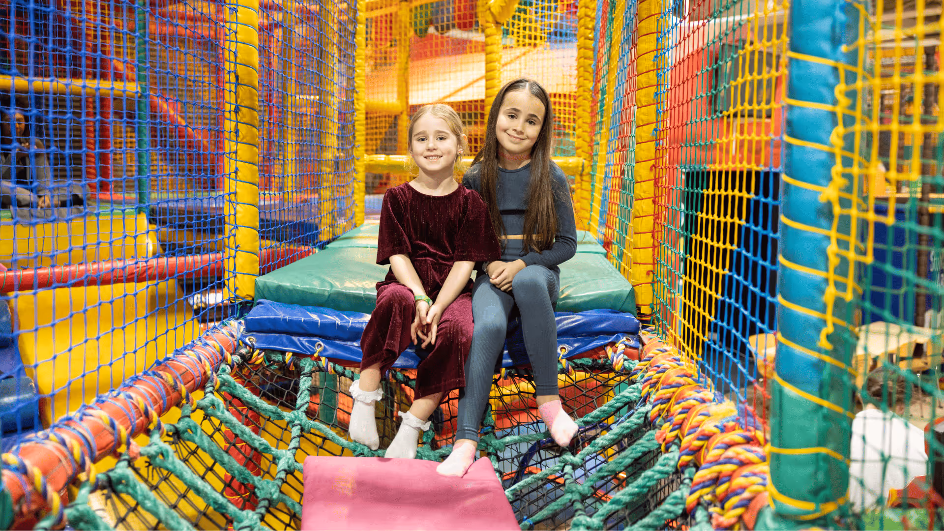 Two girls sitting in colorful indoor playground with netted play structure