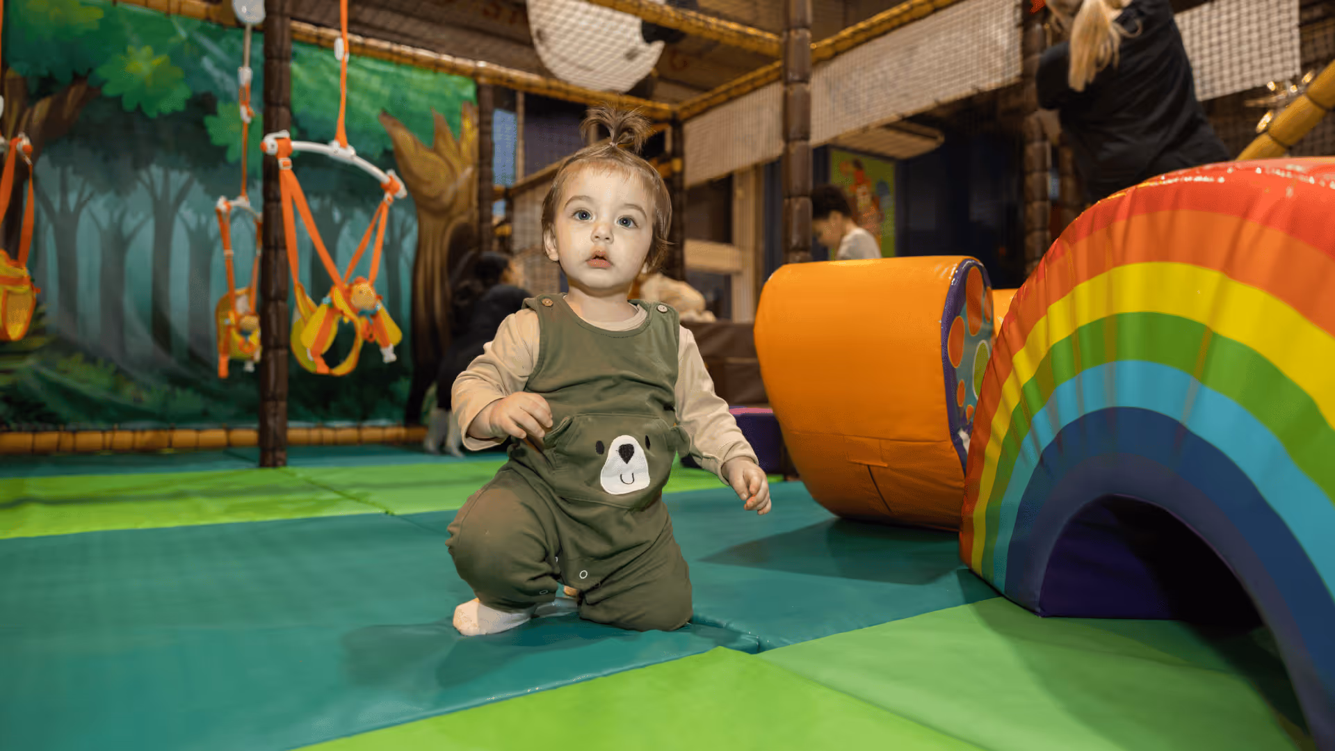 Baby sitting on colorful playmat near rainbow play structure in indoor playground
