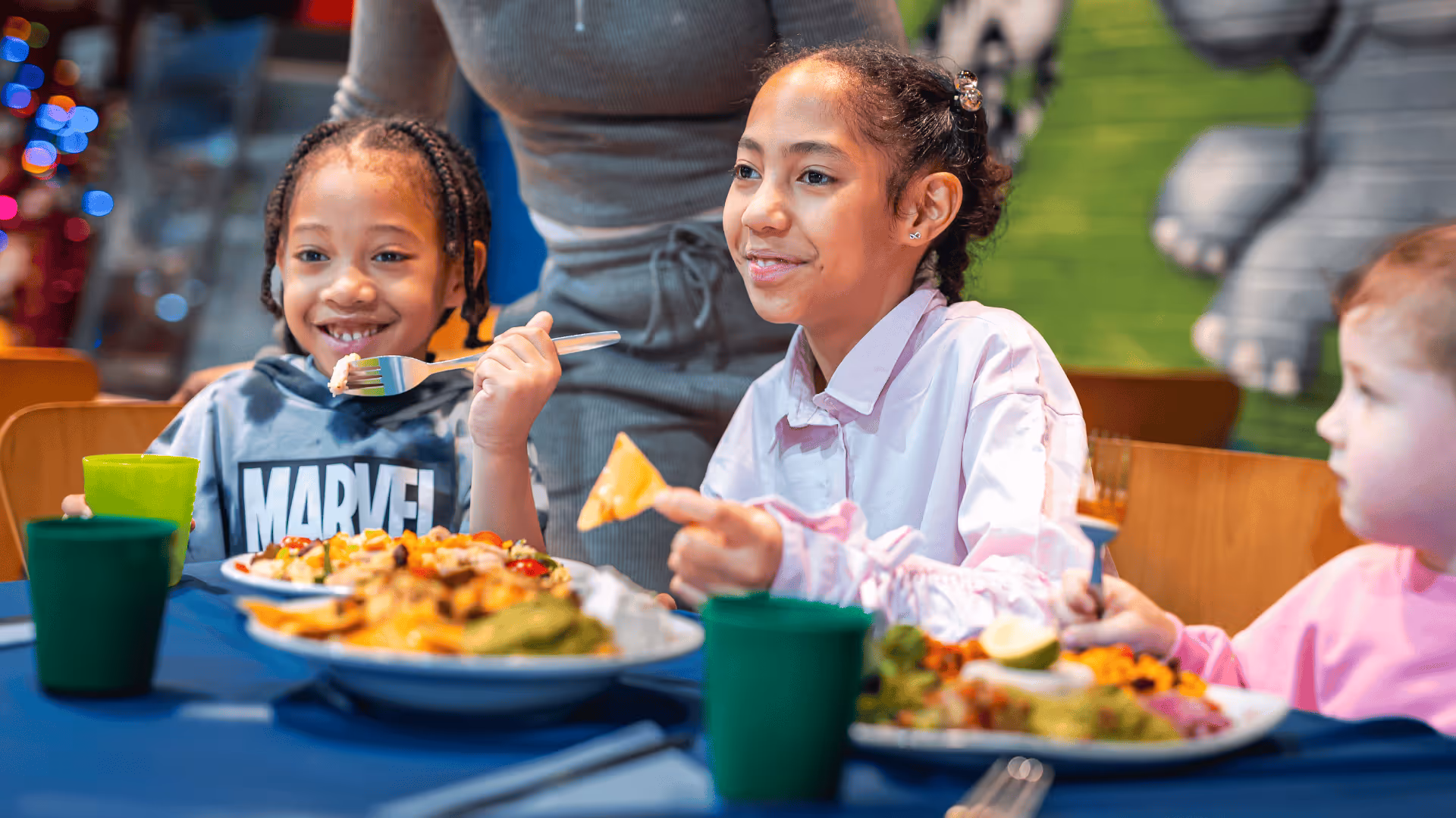 Children smiling and eating colorful meals at a table with green cups