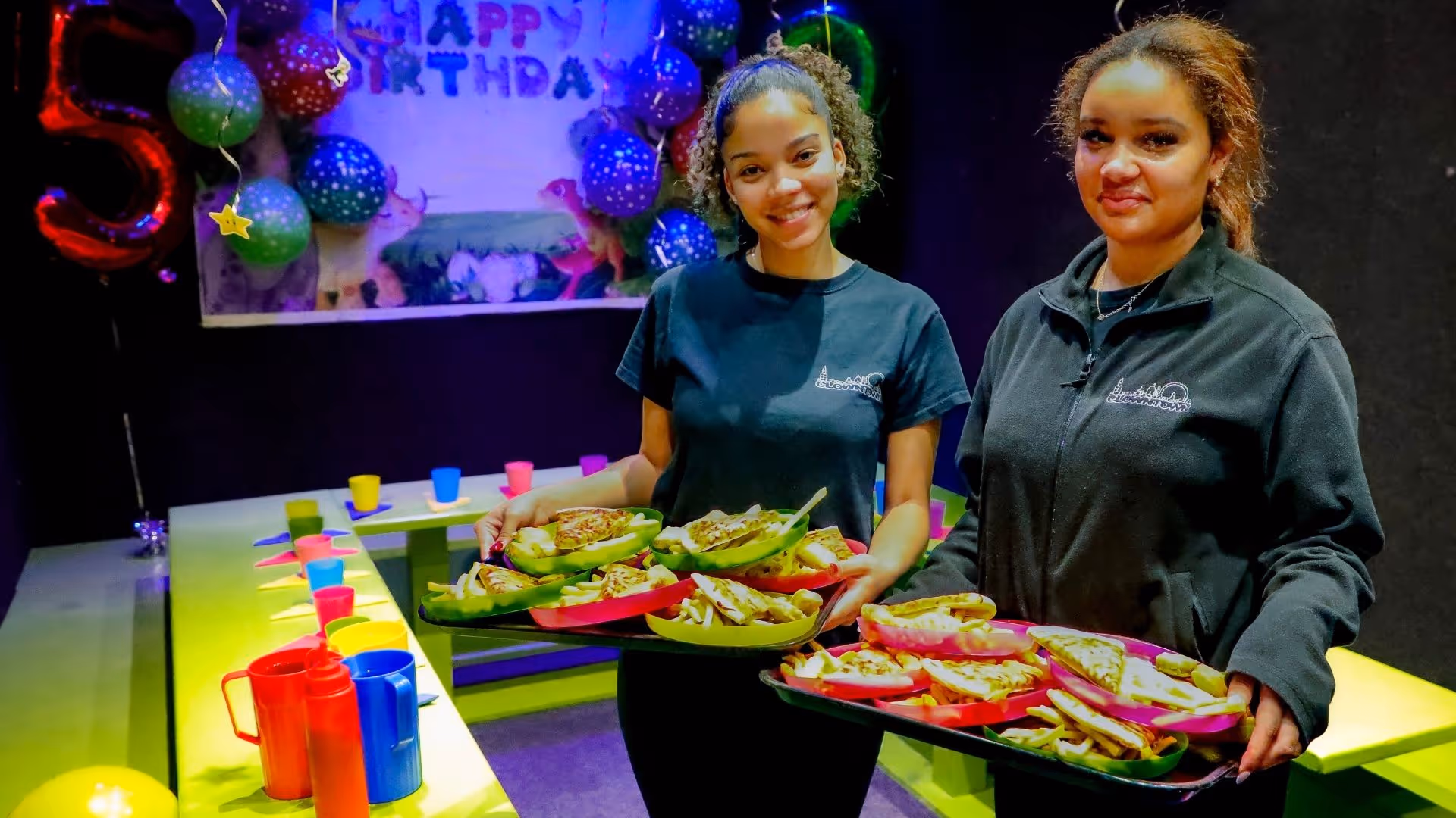 Staff serving colorful birthday party food with balloons in background