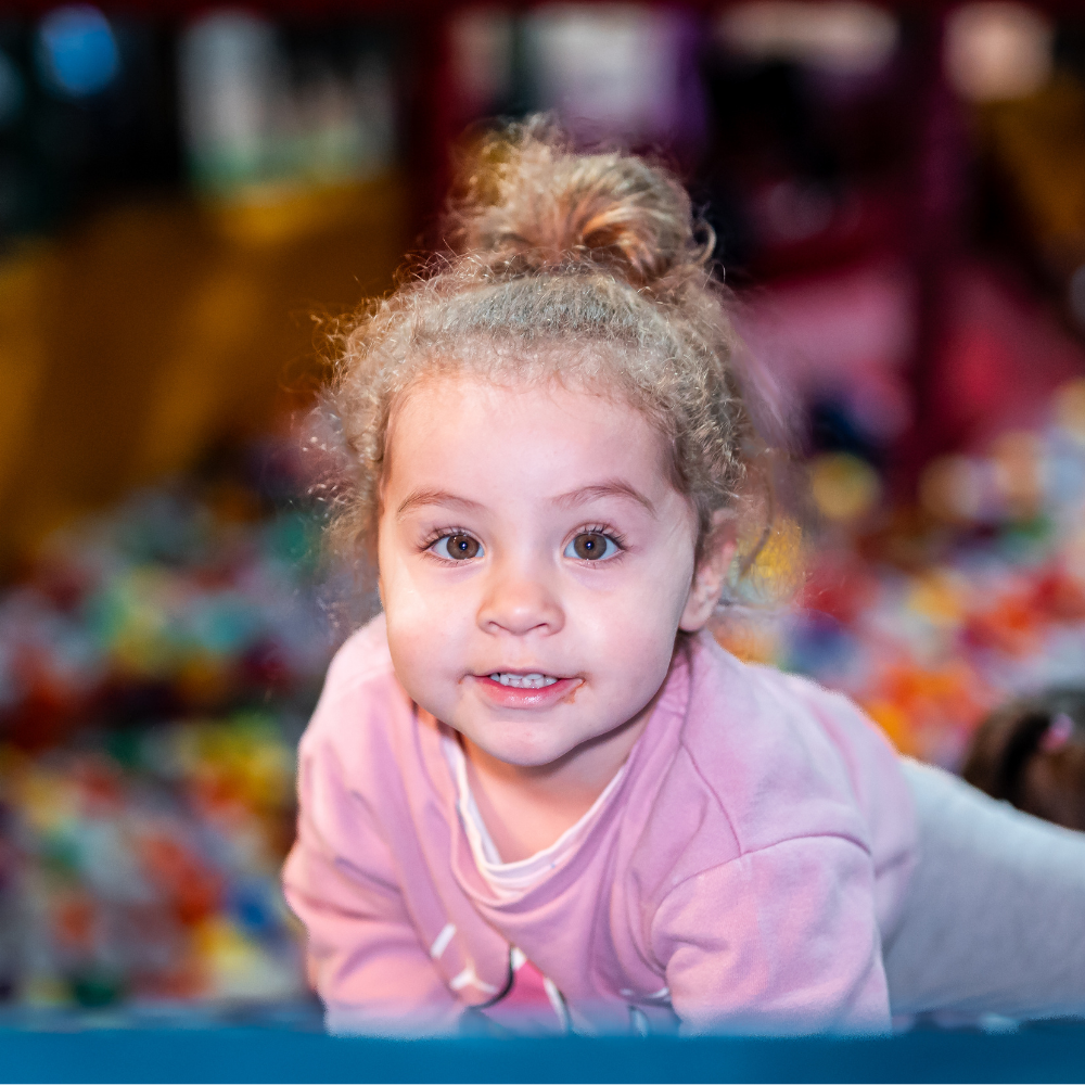 A girl smiling towards the camera in a ball pool in a soft play frame