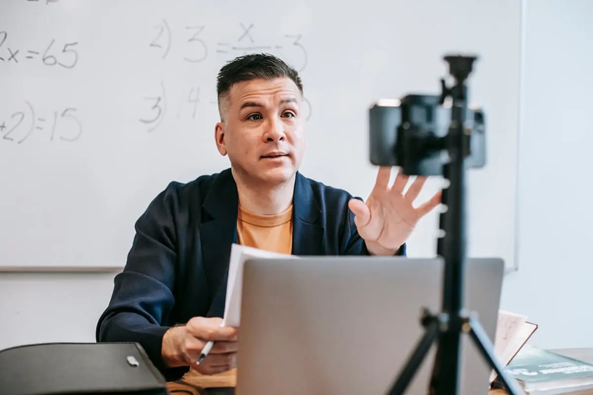 An online tutor at a desk with a laptop and whiteboard, teaching a math class via phone and computer.