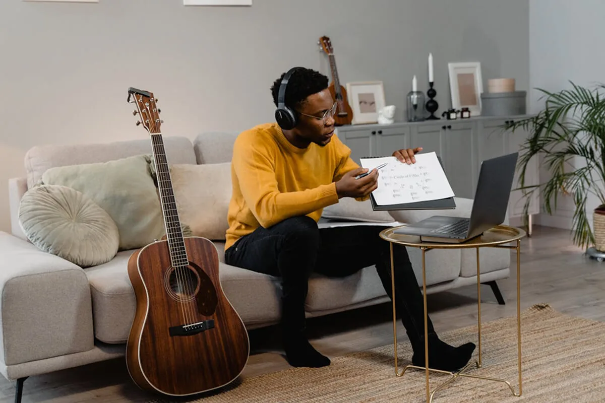 A guitarist on a sofa with a laptop and guitar, providing online guitar instruction to students from his living room.