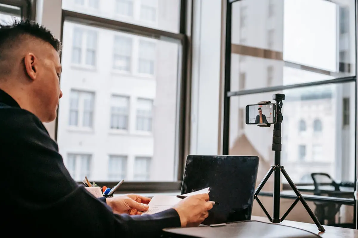 An online tutor sitting at a desk with a laptop and camera, recording a course video for his students.