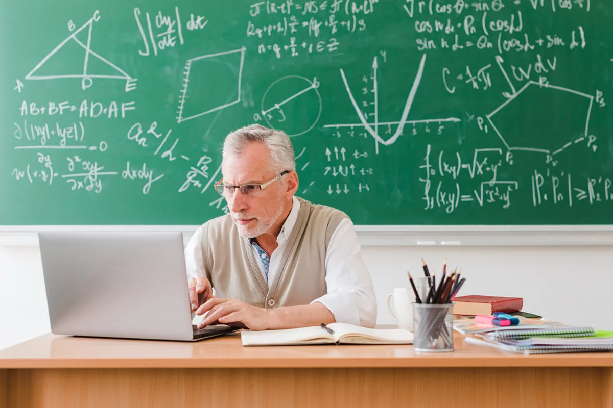 A math tutor conducting an online class through his laptop, a blackboard behind him filled with math equations.