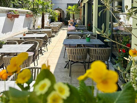 Sonnige Terrasse mit Tischen, Stühlen und Blumen, zwei Personen sitzen am Ende.