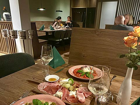 Restauranttisch mit Teller, Weingläsern, Rosen in Vase und Personen im Hintergrund.