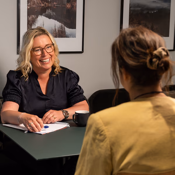 Fyra leende personer sitter på stolar i ett mötesrum medan en kvinna i förgrunden håller ett papper.
