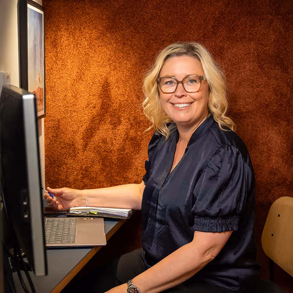 Leende kvinna med glasögon sitter vid skrivbord med laptop och bildskärm i ett kontor.