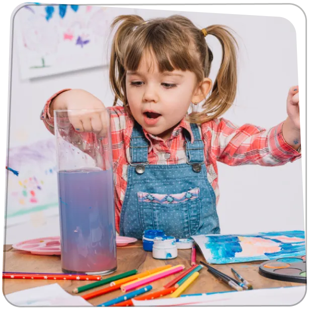 Kislány festékes asztalnál ül és önfeledten keveri a színeket, alkotás közben.