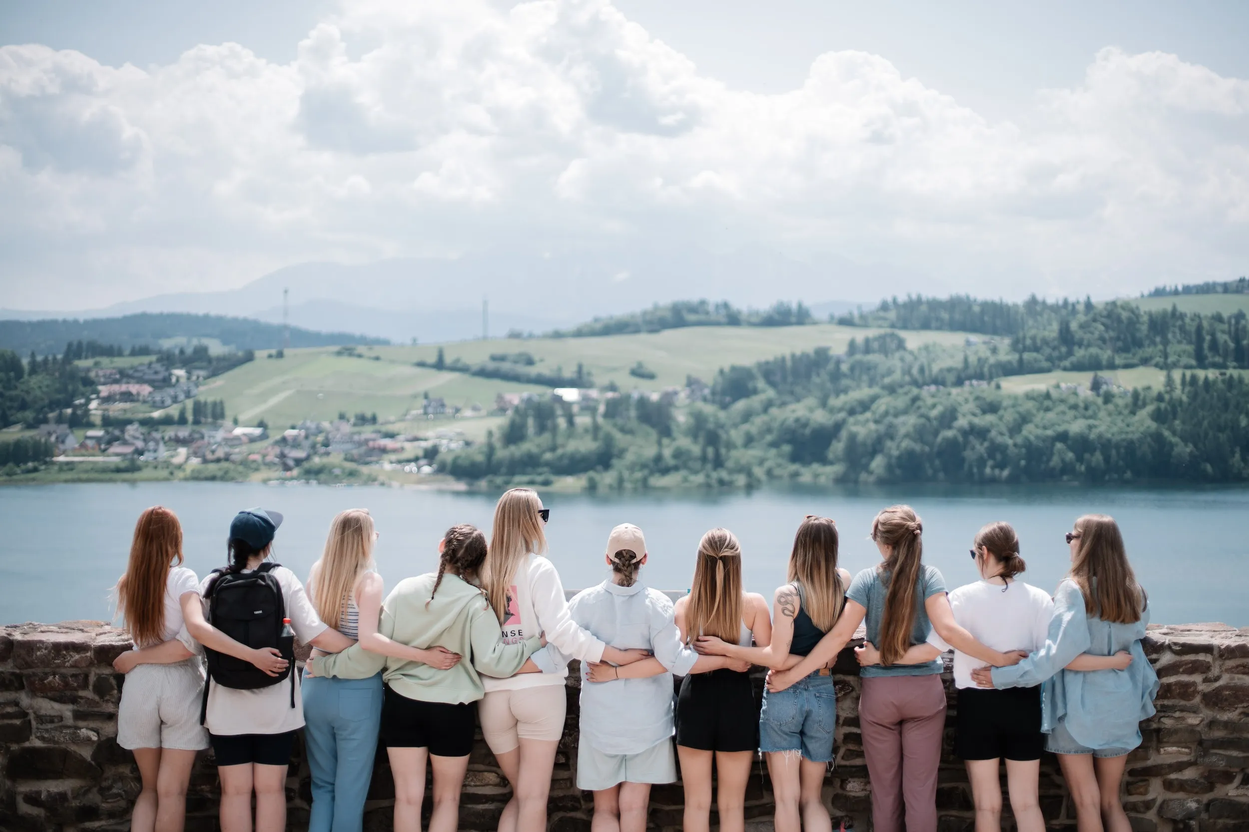 An image of 11 women standing backwards to the camera. They're looking at the lake in sunny day. They're holding their hands together behind the spines