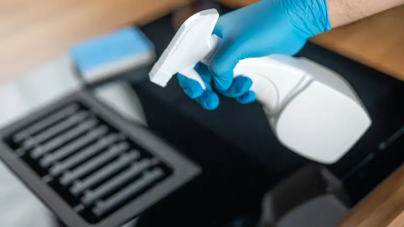 Hand in blauen Handschuhen hält eine Sprühflasche über einem Ceranfeld