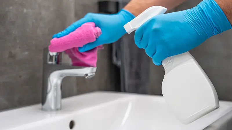 Ein Waschbecken wird geputzt, zwei Hände halten jeweils eine Sprühflasche und ein Tuch
