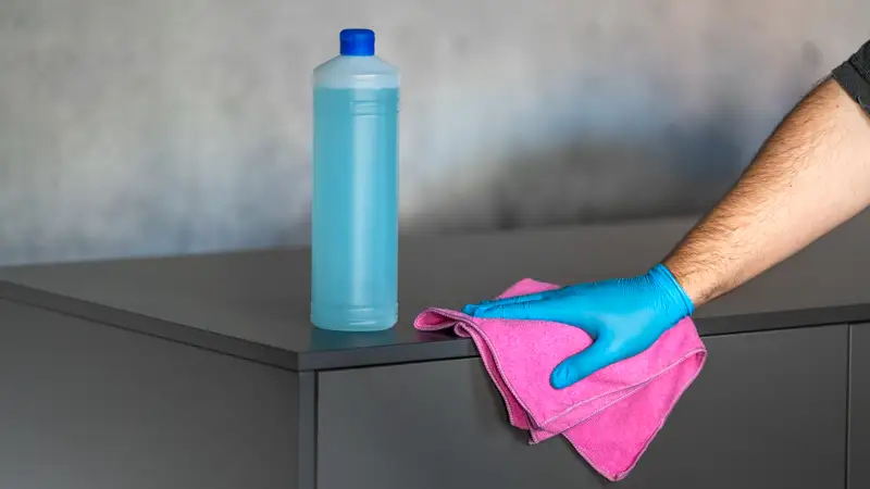 Eine Hand mit blauem Handschuh putzt ein Möbelstück mit einer Flasche im Hintergrund mit blauer Flüssigkeit