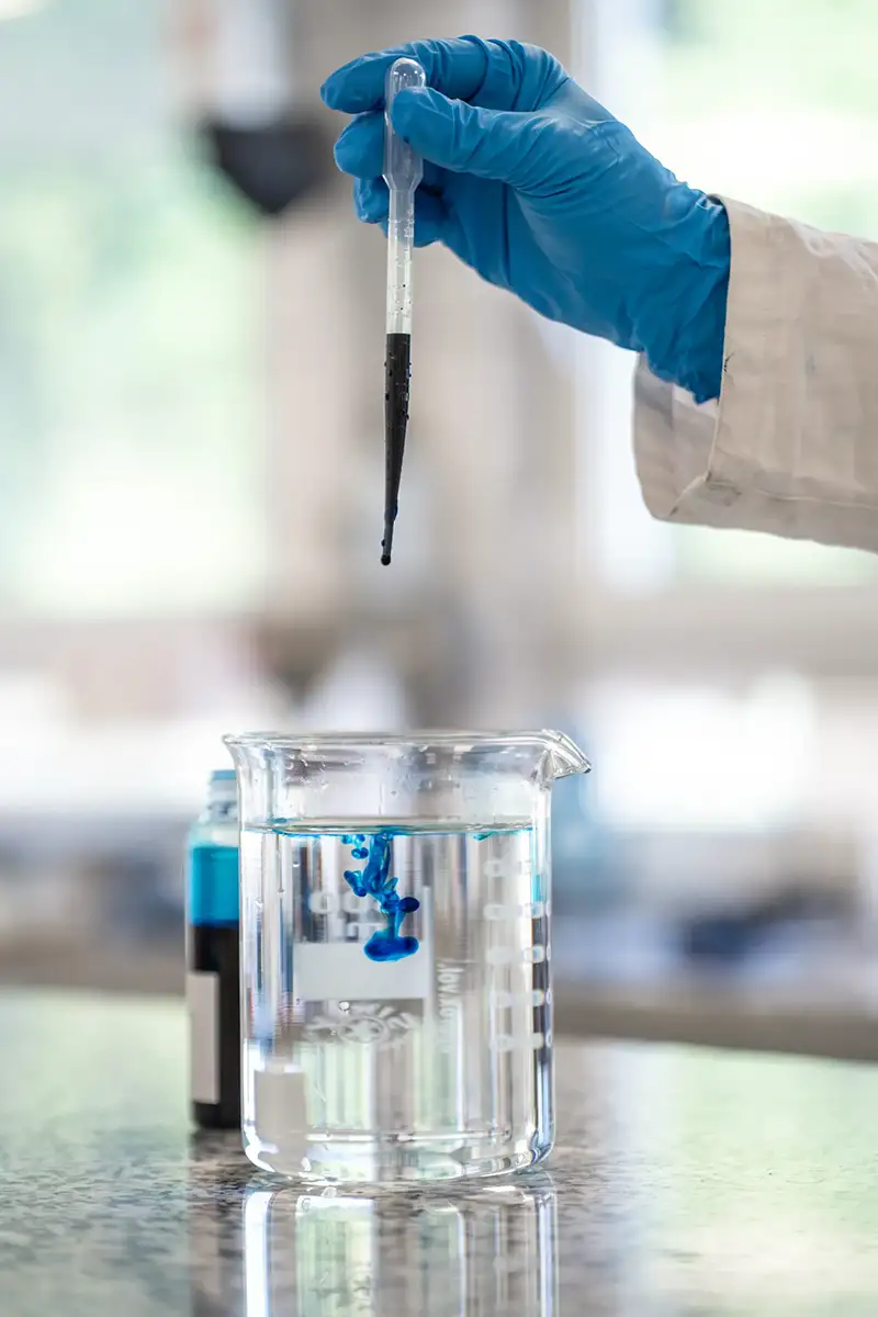 Eine Hand lässt einen Tropfen blauer Flüssigkeit aus einer Pipette in ein Glas fallen