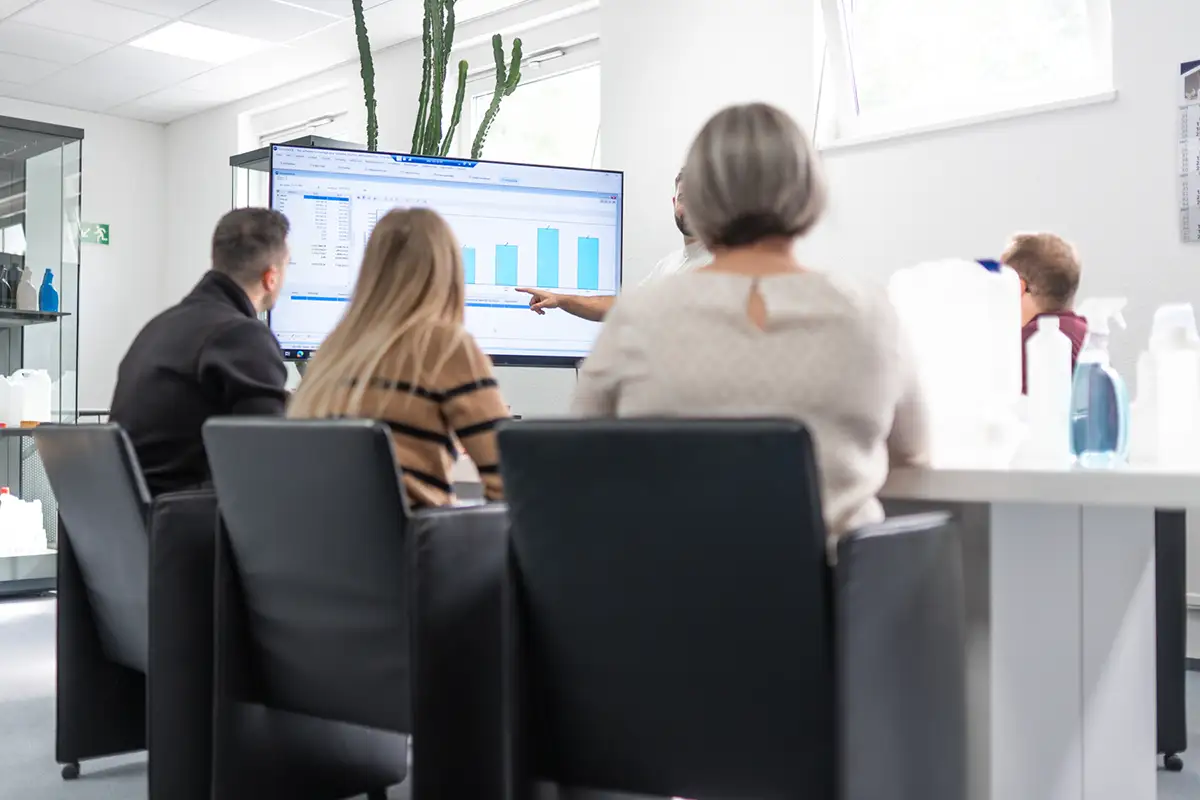Mitarbeitermeeting an einem Tisch, ein Mitarbeiter präsentiert an einem Fernseher