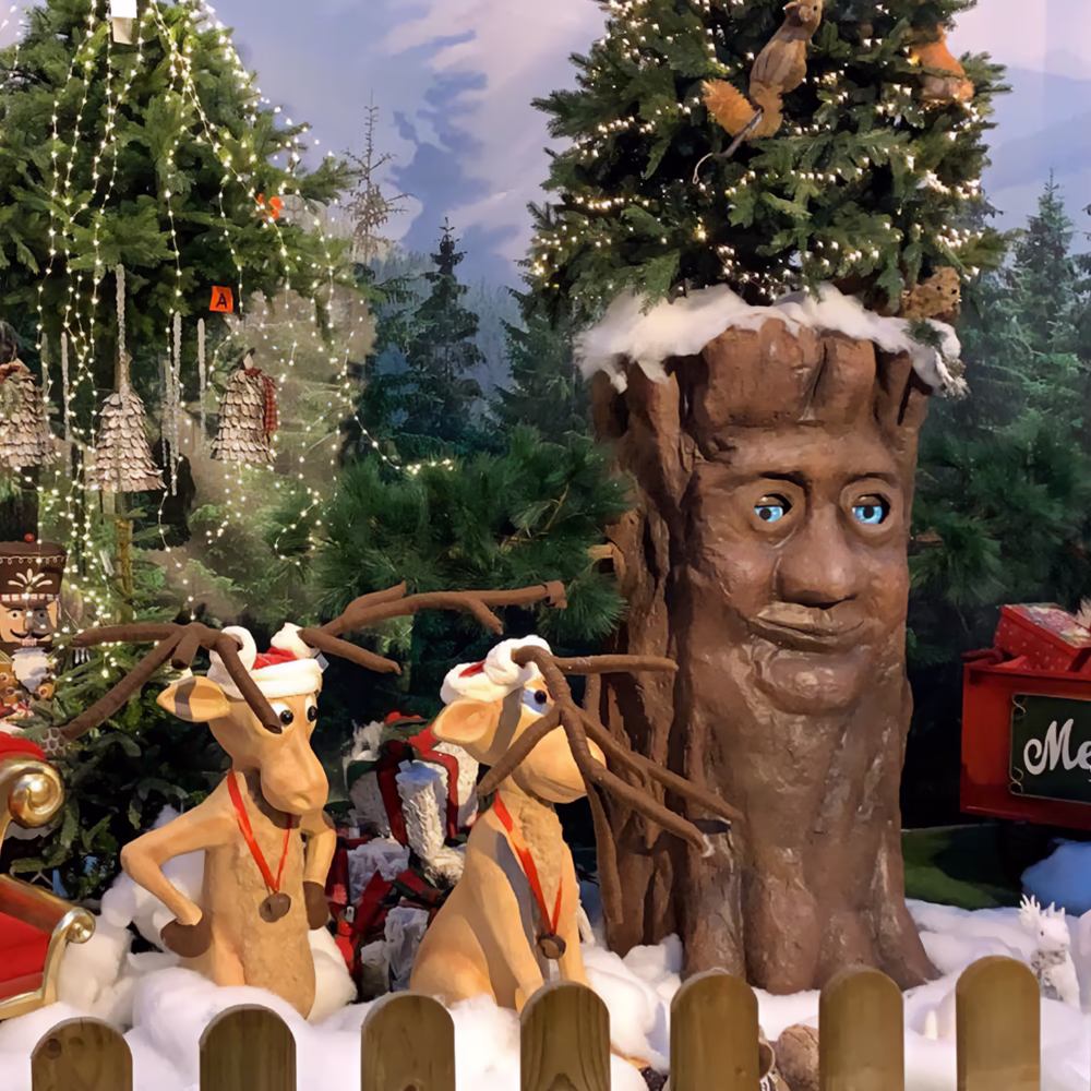 Scena natalizia con renna in giacca rossa a quadri seduta su slitta dorata, due renne con corna davanti alla slitta, albero animatronico con volto umano e corona verde. Sfondo con abeti illuminati, neve artificiale e staccionata di legno