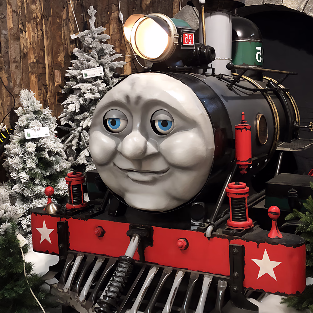Locomotiva animatronica nera con volto sorridente e occhi blu, dettagli rossi con stelle bianche, faro acceso. Tunnel di pietra ad arco, alberi di Natale innevati e lanterne rosse come decorazioni