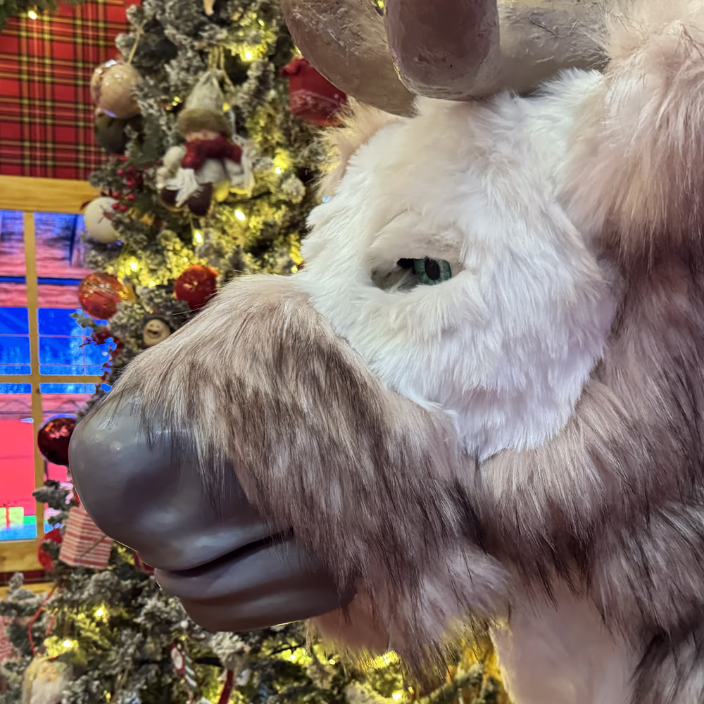 Renna animatronica con pelo marrone e bianco, naso nero prominente, in ambiente natalizio. Sfondo con tessuto a quadri rossi, albero di Natale decorato con luci dorate e cappotto rosso di Babbo Natale appeso a sinistra