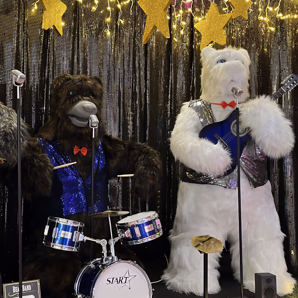 Tre orsi animatronici formano una band: orso grigio con trombone e abito luccicante a sinistra, orso con batteria e vestito blu al centro, orso bianco con chitarra elettrica blu a destra. Sfondo dorato con tende metalliche e stelle, tutti indossano papillo