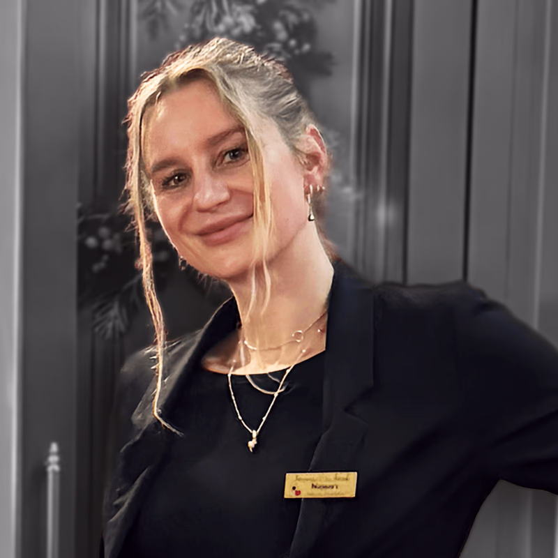 Blonde woman with tied hair and name badge, black top and gold necklaces, smiles leaning against a gray door.