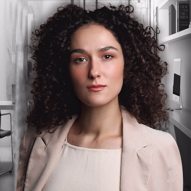 Woman with dark voluminous curly hair, light beige jacket, photographed in modern kitchen with appliances and white shelves.