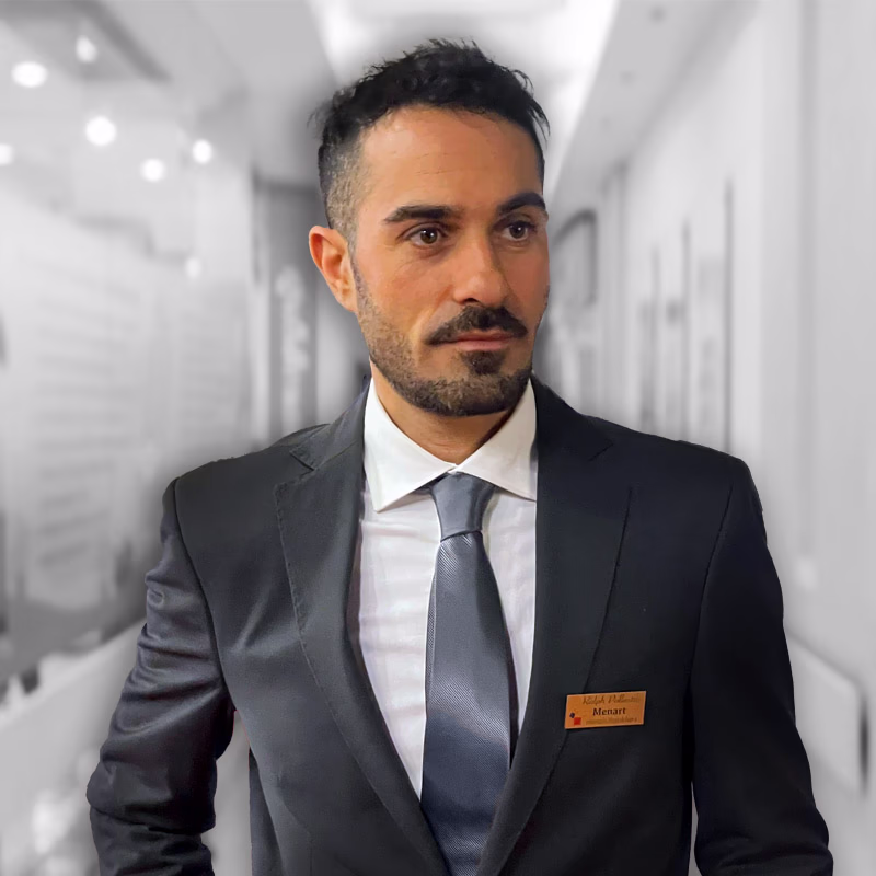 Professional man with beard and name badge, in dark suit with gray tie, in modern illuminated hallway.