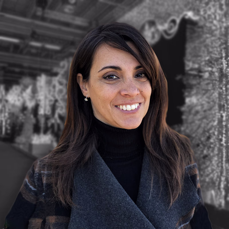 Smiling woman with long brown hair, black sweater and gray checked jacket, photographed in an outdoor urban context.