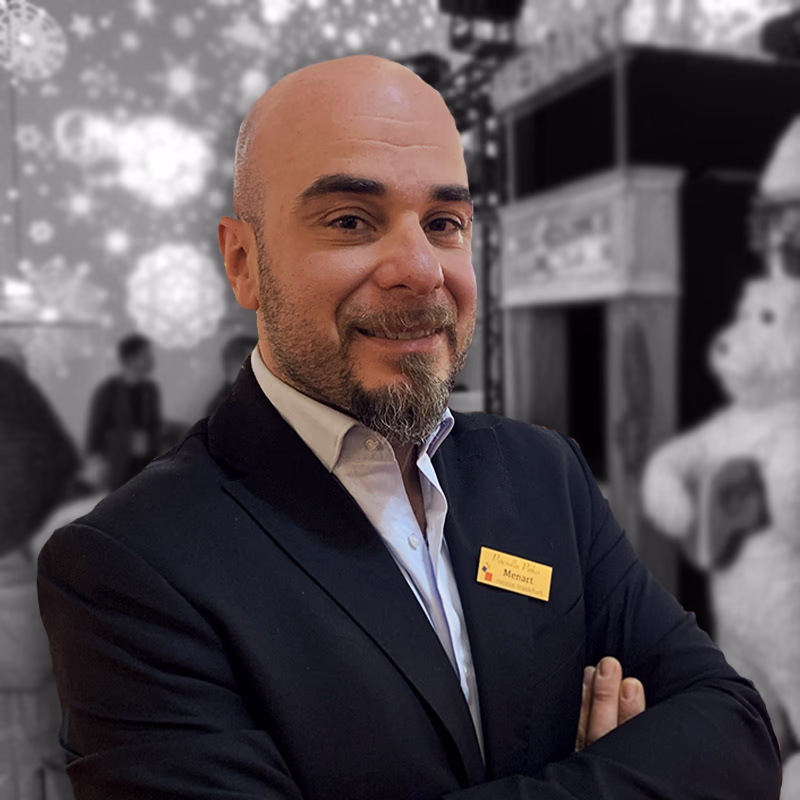 Bald man with beard and name badge, in a dark jacket, smiles with folded arms in an elegant business environment.