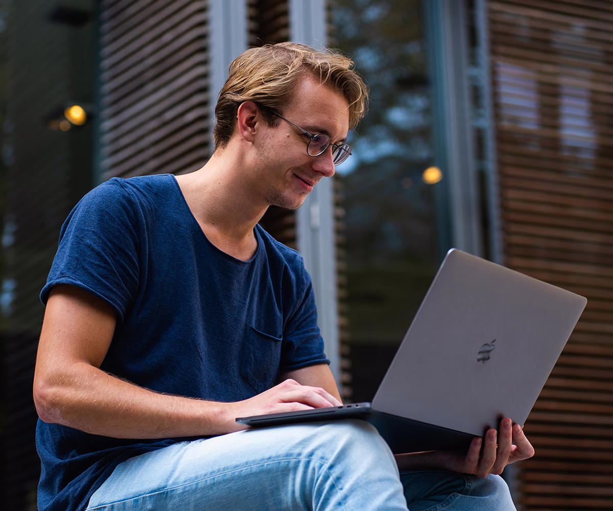 A young man with blond hair and glasses wearing a blue t-shirt is sitting outside while working on a laptop. He is focused on the screen as he types, with Venetian blinds and warm evening lighting visible in the background. The environment appears to be a covered outdoor area or patio, creating a relaxed atmosphere for remote work.