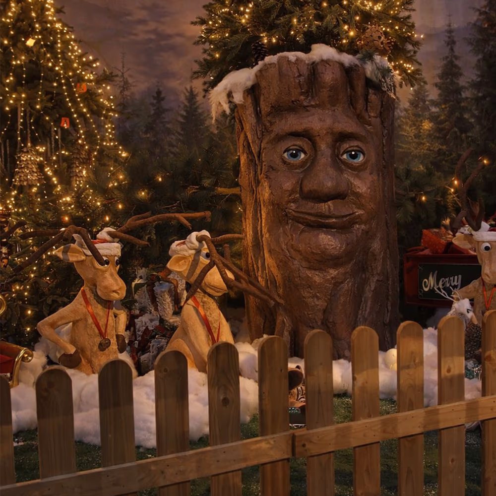 Albero di Natale con volto umano in mezzo a renne di decorazione con luci festive e recinzione di legno.
