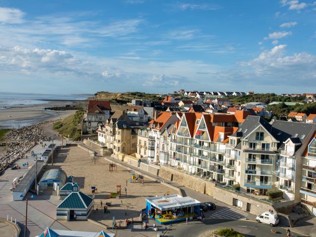Vue aérienne d'une plage bordée d'immeubles avec toits rouges et blancs, une promenade piétonne et une mer sous un ciel partiellement nuageux de la côte d'Opale.