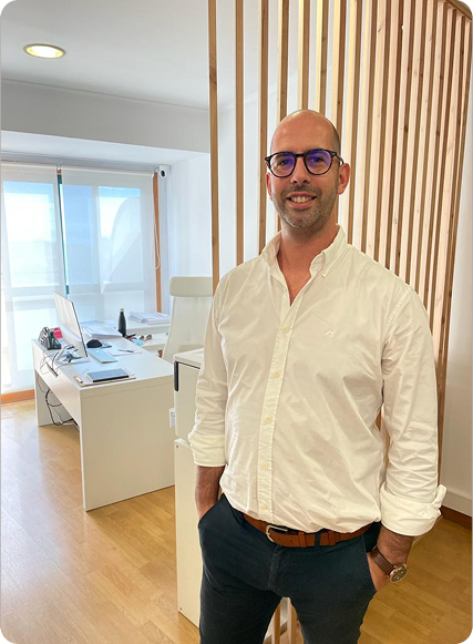 Fotografia de corpo de Miguel Henriques, arquiteto e gerente da MHArquiteto, que posa no seu escritório, vestido de camisa branca e calças escuras com um cinto castanho. É um homem careca, com óculos e sorridente. Pousa com as mãos nos bolsos, sobre uma divisória de ripados de madeira com  uma secretária e janelas atrás.