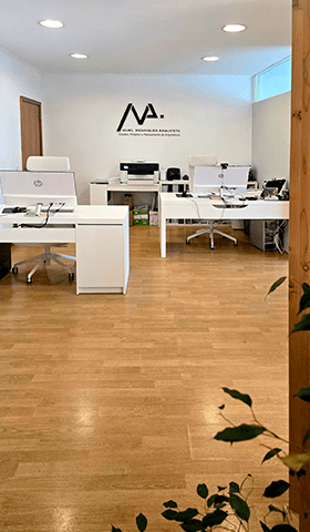 Fotografia do interior do escritório de MHArquiteto. Paredes brancas com o logo da MHArquiteto colocado sobre a parede de fundo, em grande. Soalho de pinho, de tom claro e duas secretárias com monitores por cima.