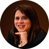 Mulher com cabelo escuro comprido, a sorrir de forma ligeira, apoiando o queixo na mão. Retrato em plano aproximado, com fundo neutro escuro.